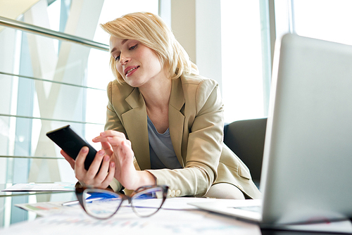 Portrait shot of pretty blond-haired manager texting with friend on smartphone while distracted from work, interior of modern office with panoramic windows on background