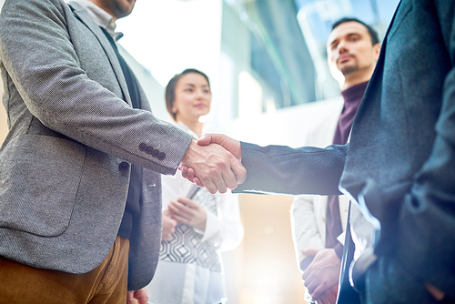 Mid-section of two successful business people shaking hands after beneficial deal, people in background
