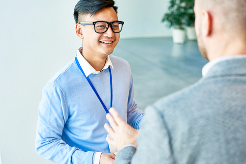 High angle portrait of cheerful Asian businessman wearing glasses smiling happily while talking to colleague at work