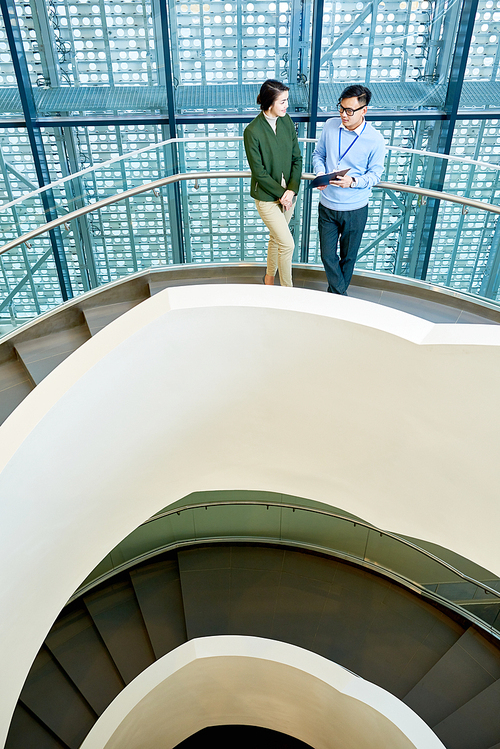 High angle view of pretty young manager standing on spiral concrete staircase and listening to her male colleague with interest while discussing start-up project