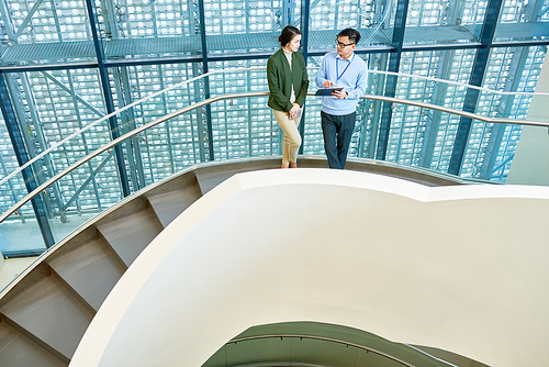 Attractive young manager and her male colleague standing on spiral concrete staircase of modern office building and analyzing work results, high angle view