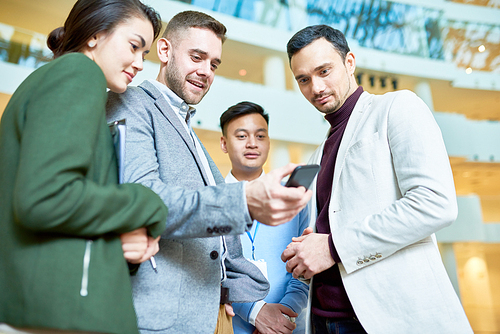 Low angle portrait of modern businessman holding smartphone showing something to group of colleagues standing in hall of office building