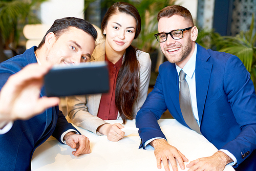 Portrait of three young business people taking group selfie via smartphone while sitting at cafe table during coffee break