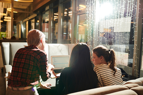 Rear view of young female friends sitting in cafe together and communicating online using laptop