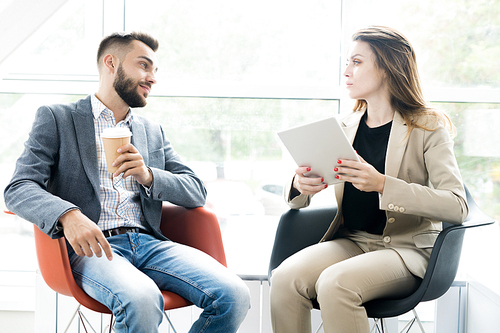 Portrait of two modern business people, man and woman, sitting in designer chairs and talking to each other in sunlit room