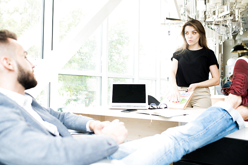 Portrait of young businesswoman discussing project with chill colleague sitting at meeting table with feet up
