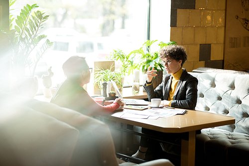 Serious intelligent curly-haired business lady in jacket sitting at table with papers and gesturing hand while sharing her opinion with colleague at meeting in loft restaurant.
