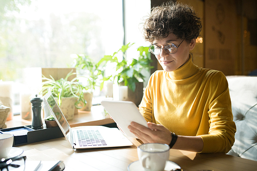 Content pensive curly-haired female freelancer in glasses sitting at table in modern cafe and using digital tablet while analyzing online information