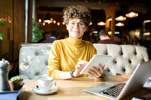 Smiling attractive hipster lady with short curly hair sitting at table and using digital tablet while working in restaurant, she 