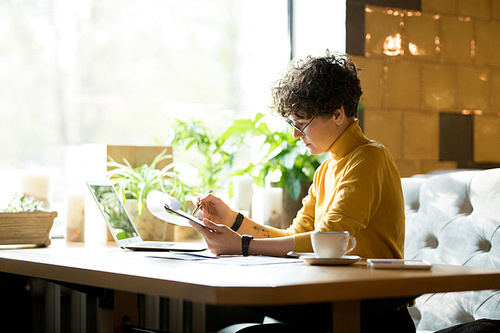 Serious concentrated young businesswoman with curly hair sitting at table and viewing report while finding mistakes in modern cafe