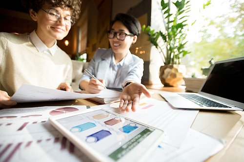 Close-up of confident attractive multiethnic business ladies in casual clothing sitting at table and analyzing scheme on tablet while planning project together