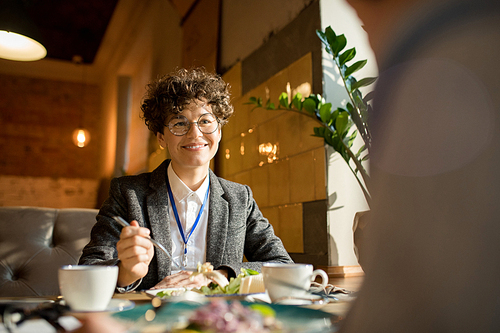 Happy content attractive curly-haired businesswoman with badge sitting at table and eating salad while talking to colleague over lunch