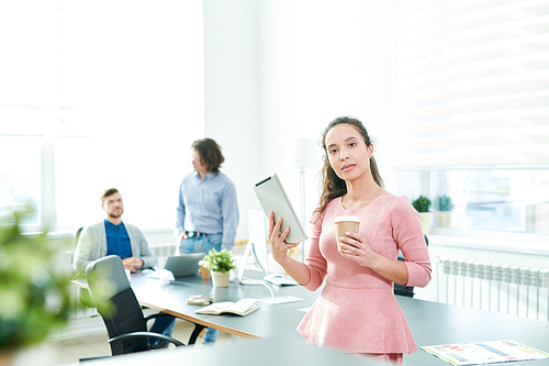 Serious confident beautiful young Hispanic female manager with modern device and coffee cup  while her colleagues discussing business plan in background