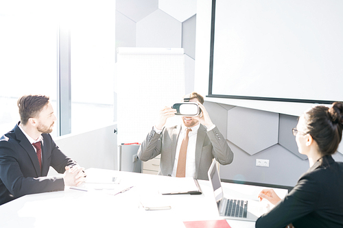Portrait of contemporary businessman enjoying  VR while sitting at meeting table in conference room with colleagues, copy space