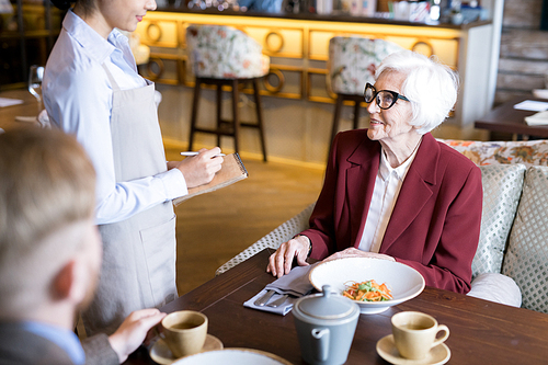 Elegant senior woman making an order to waitress while sitting at the table and eating together with young man