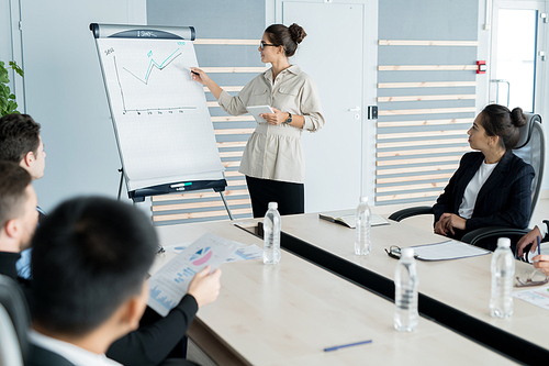 Young sales coach with hair bun wearing glasses holding presentation and explaining graph to business people at training class