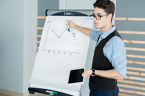Serious smart young male manager in glasses standing at whiteboard and pointing at graph while presenting sales report