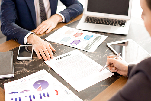 Close-up of business lady reading contract and preparing to sign it after negotiation with financial specialist, they sitting at table full of analytical papers and modern devices