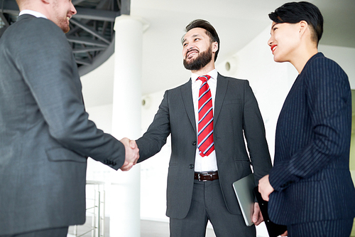 Smiling bearded entrepreneur greeting his business partner with firm handshake while standing at spacious office lobby, pretty assistant manager looking at them with wide smile