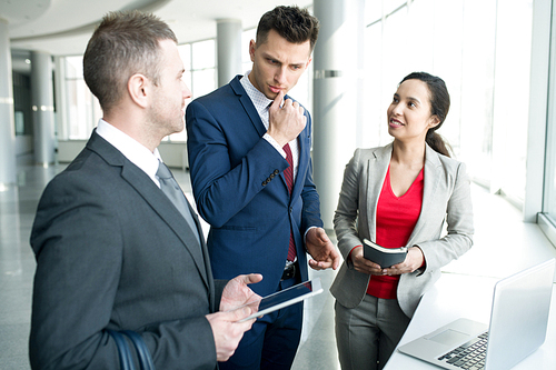 Group of modern business people discussing work plans and strategy standing by window in modern office, focus on frowning young man deep in thought