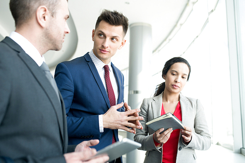 Group of modern business people discussing work plans standing by window in modern office, focus on handsome young man explaining strategy to partners