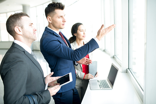 Waist up portrait of group of modern business people discussing work plans standing by window in modern office, focus on handsome young man pointing to window