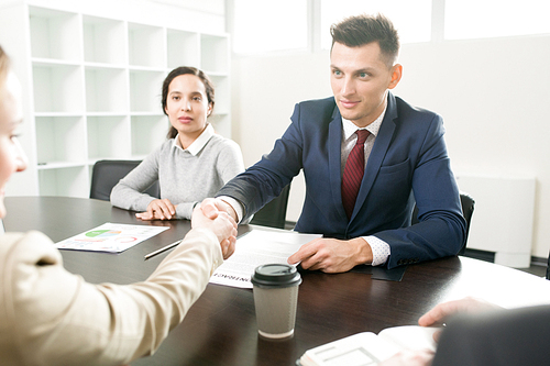 Content confident handsome business representative making handshake with new business partner while sitting with assistant at table during meeting