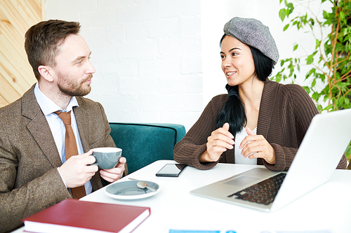 Portrait of trendy young businesswoman talking to colleague meeting during coffee break  while sitting at table in cafe and using laptop