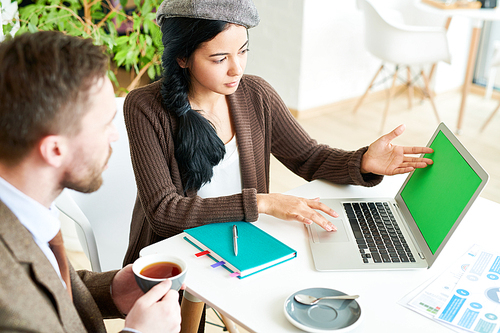 Portrait of pretty modern woman pointing at laptop with green screen during meeting in cafe with partner or client, discussing possible cooperation and presenting services