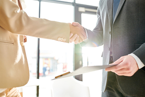 Mid section side view of two unrecognizable  business people shaking hands standing against window in sunlight and holding documents after successful deal