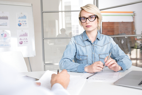 Portrait of young blonde businesswoman wearing casual clothes and glasses sitting at meeting table in office  listening to partner with serious face expression, copy space