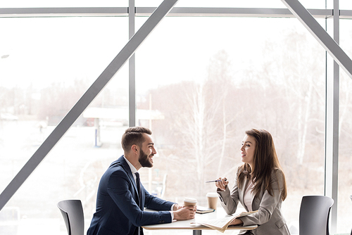 Side view portrait of two cheerful business people, man and woman, drinking coffee at table in cafe sitting at big window, copy space