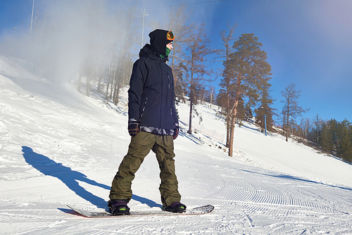 Full length portrait of modern  young man riding snowboard at ski resort piste standing against clear blue sky, copy space