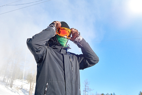 Low angle portrait of modern young man wearing snowboarding gear and face mask ready to go freestyle riding downhill in snow covered mountains, copy space