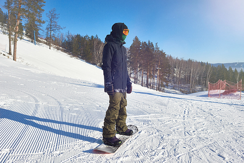 Full length portrait of modern  young man riding snowboard at snow covered ski resort piste, copy space