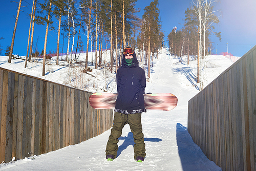 Full length portrait of modern young man posing with snowboard and  standing between wooden fences at ski resort, copy space