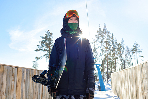 Low angle portrait of modern young man holding snowboard and looking away with determination standing against clear blue sky, lens flare, copy space