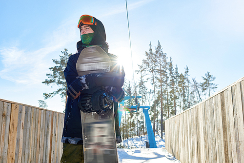 Low angle portrait of modern snowboarder and looking away with determination standing against clear blue sky, lens flare, copy space