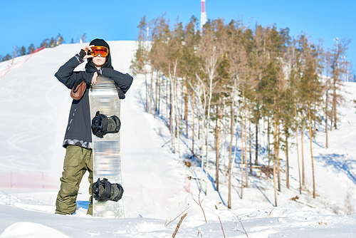 Full length portrait of successful young rider posing with snowboard looking confidently away standing against mountain piste, copy space