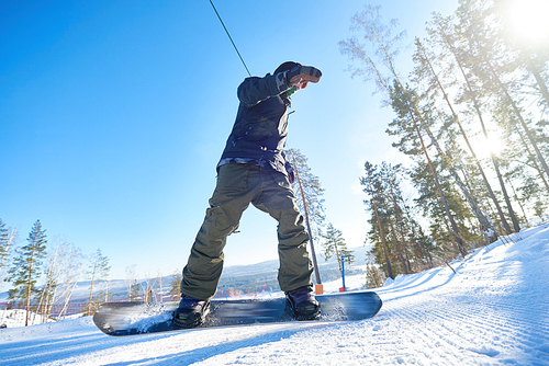 Low angle portrait of young man riding snowboard on mountain piste at ski resort in sunlight, copy space