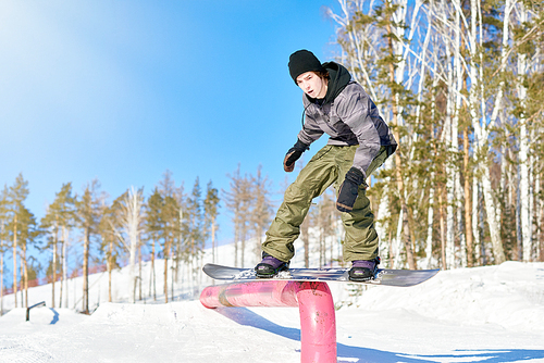 Full length action shot of young man performing snowboarding stunt sliding down metal railing in sunlight at ski resort, copy space