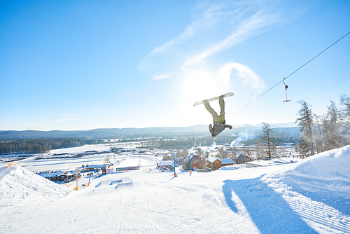 Full length action shot of young man performing snowboarding stunt backside flip jumping in air in sunlight at ski resort, copy space