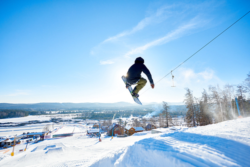 Back view action shot of young man performing snowboarding stunt jumping high in  sunlight at ski resort, copy space