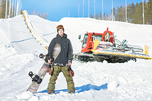 Full length portrait of modern young snowboarder posing at foot of snow covered piste with snowcat in background, copy space