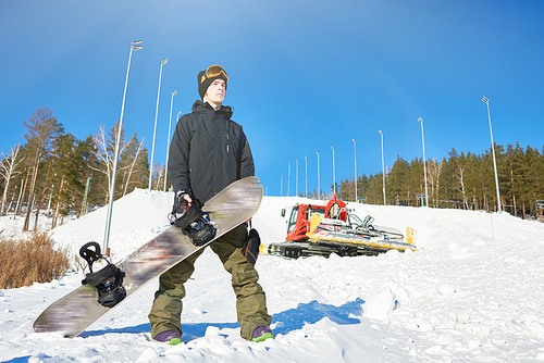 Full length portrait of modern young snowboarder posing at foot of snow covered hill with snowcat in background, copy space