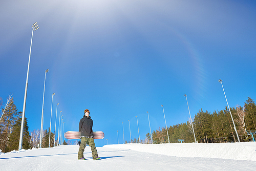 Full length wide angle portrait of modern young snowboarder posing on empty piste at ski resort against clear blue sky, copy space