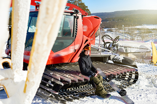 Full length portrait of modern young snowboarder sitting on caterpillar drives of snowcat at snow covered piste of ski resort, copy space