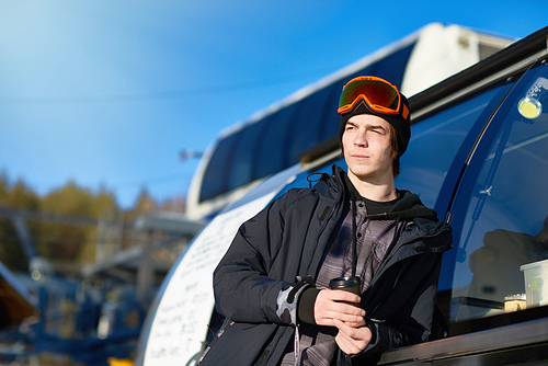 Portrait of modern young man wearing snowboarding gear relaxing leaning on truck and warming up with coffee, copy space