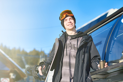 Portrait of modern young snowboarder standing by on truck and smiling happily enjoying sunny winter day at ski resort, copy space