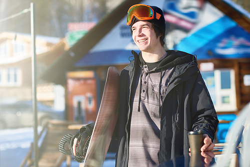 Portrait of modern young snowboarder smiling happily enjoying sunny winter day at ski resort with chalets in background, copy space